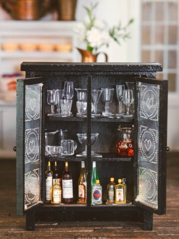 A black and metal miniature cupboard open with mini stemware and bottles inside