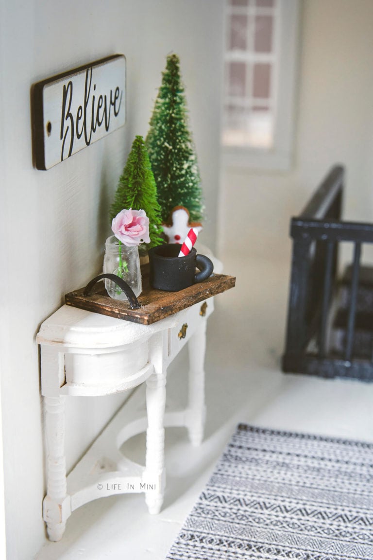 A wooden tray with a miniature mason jar, rose and small Christmas trees on a white console table