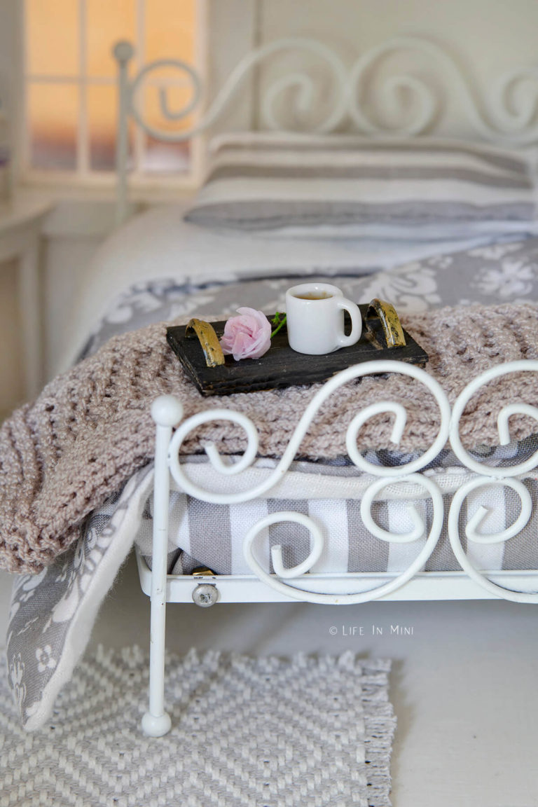 A black mini tray with gold handles with a rose and mug in it and sitting on a dollhouse bed