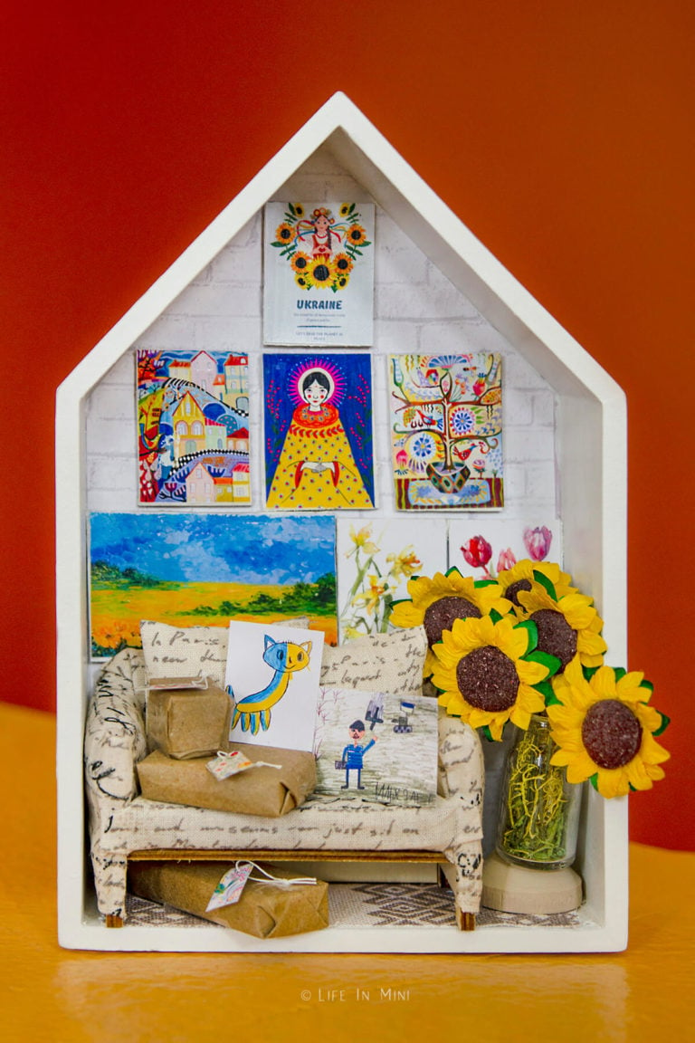 A miniature scene in a small box shaped as a house depicting a small sofa with packages on it, colorful Ukrainian artwork and sunflowers in it