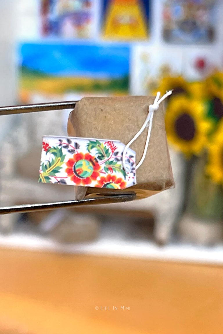 Closeup of tweezers holding a miniature package wrapped in brown paper with a miniature floral tag on it