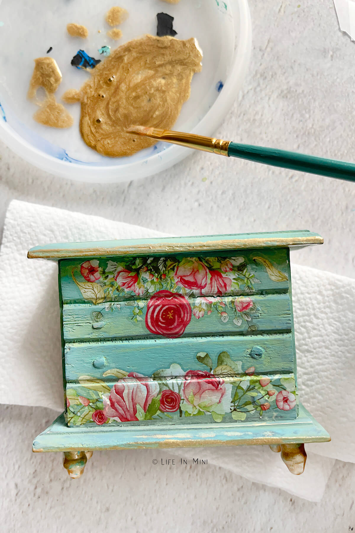 Painting gold trim over a teal dollhouse dresser with floral prints
