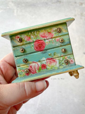 A hand holding a 1:12 teal dollhouse dresser with floral rub ons and gold touches