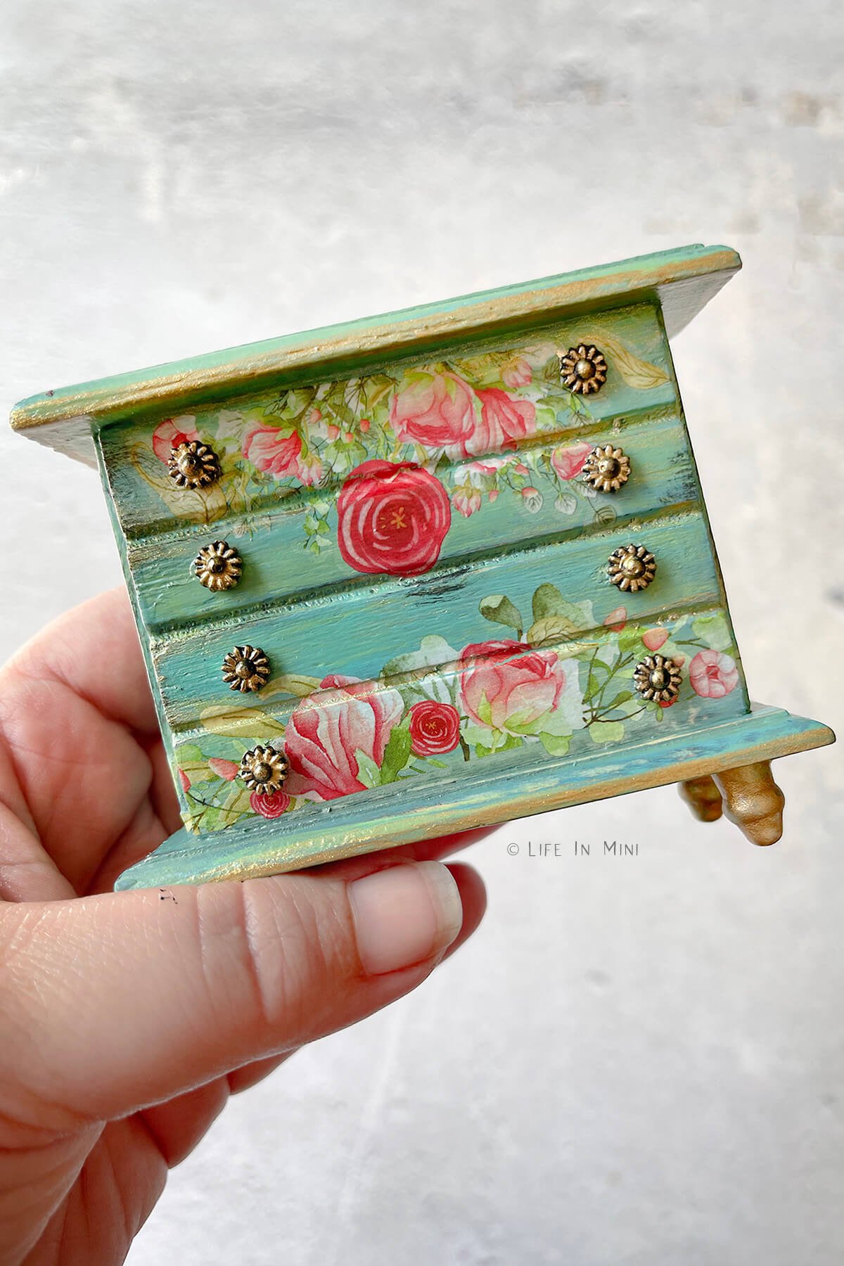 A hand holding a 1:12 teal dollhouse dresser with floral rub ons and gold touches