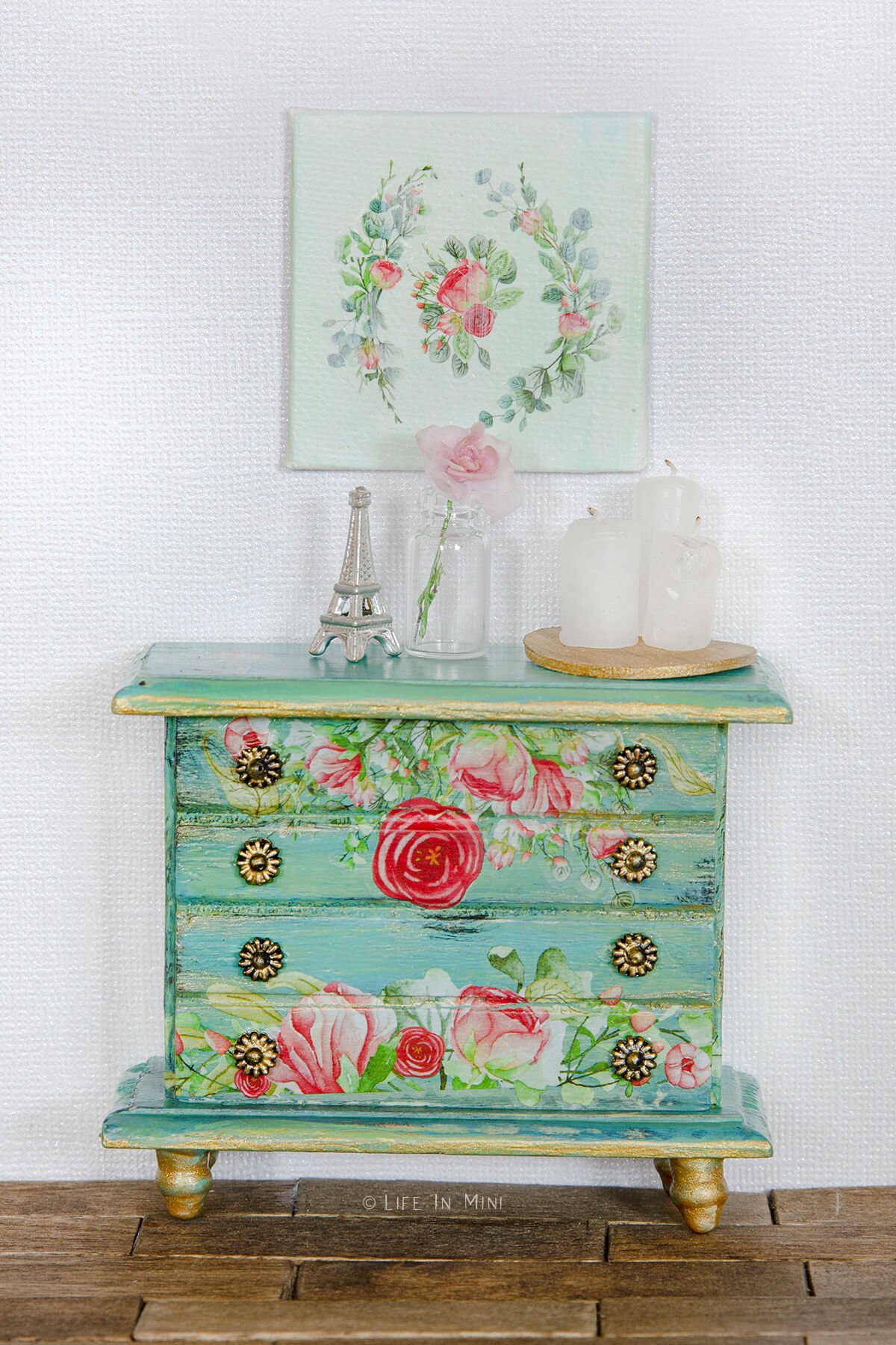 A teal dollhouse dresser with floral rub ons with miniature accessories on it