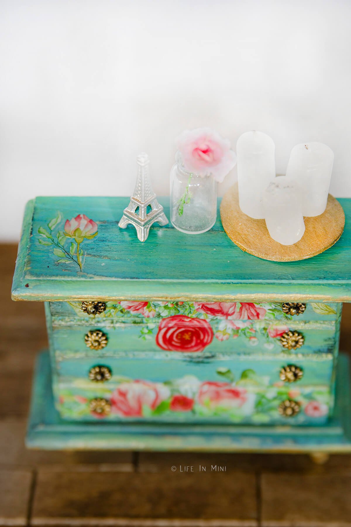 Closeup of miniature accessories sitting on top of a teal dollhouse dresser with floral rub ons