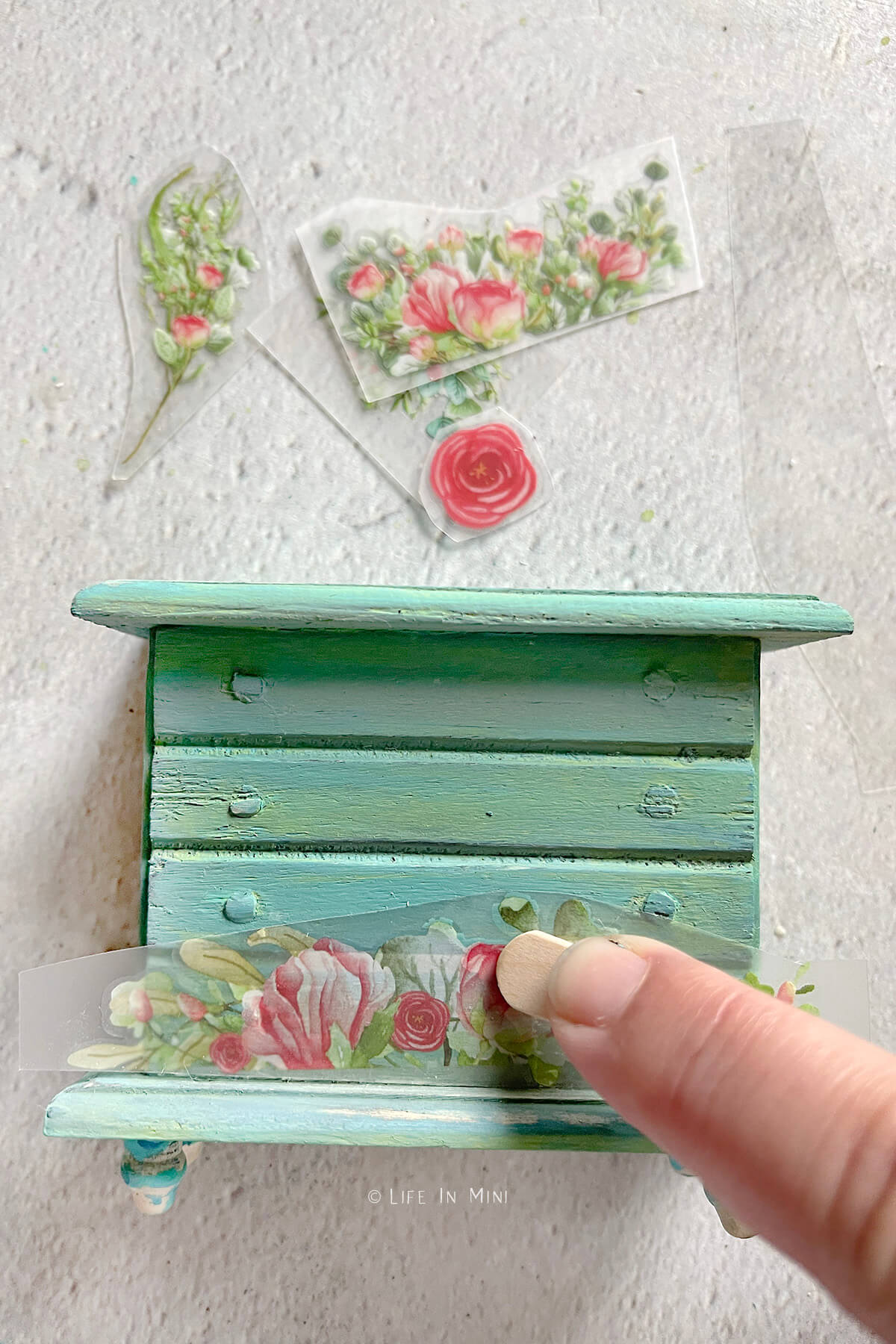 Pressing on floral rub on transfers onto a teal dollhouse dresser