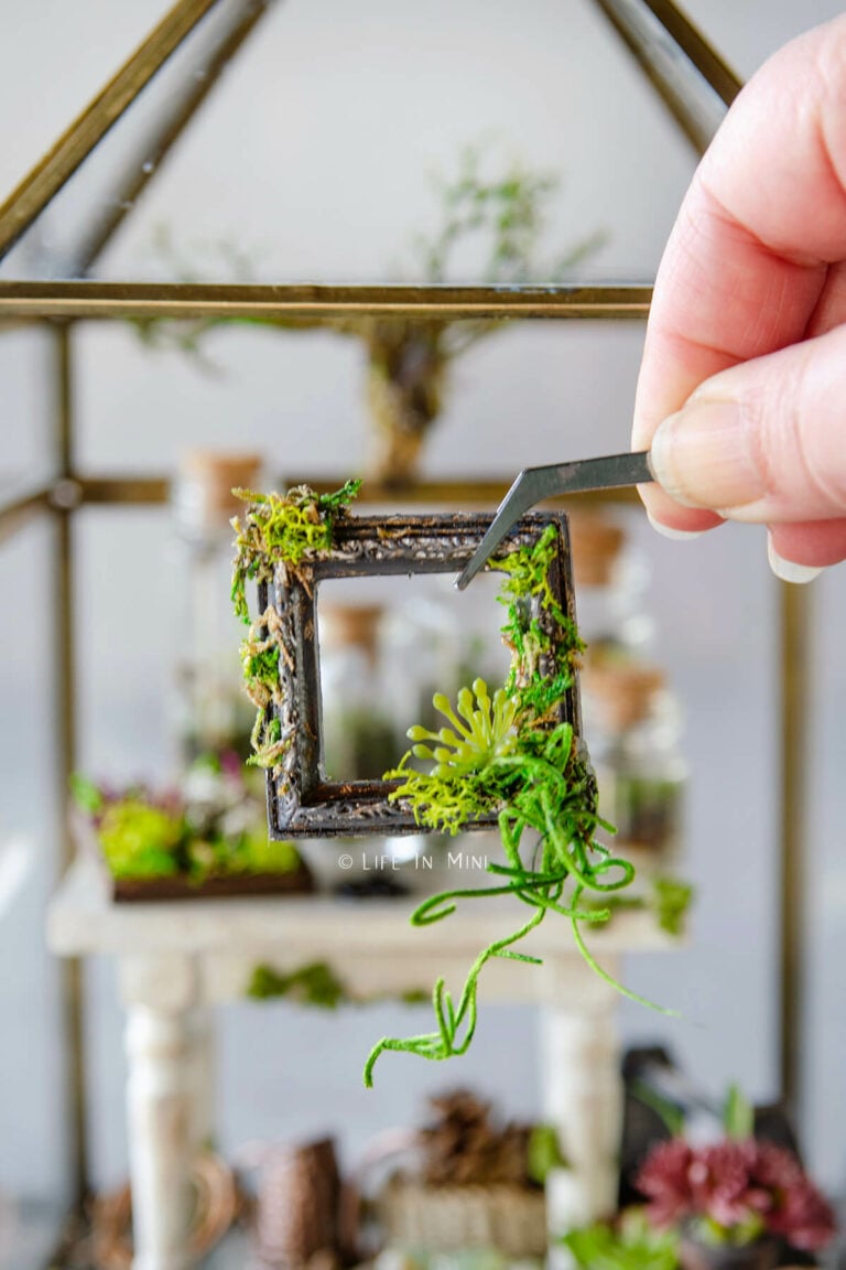 A hand holding tweezers holding a moss covered mini picture frame
