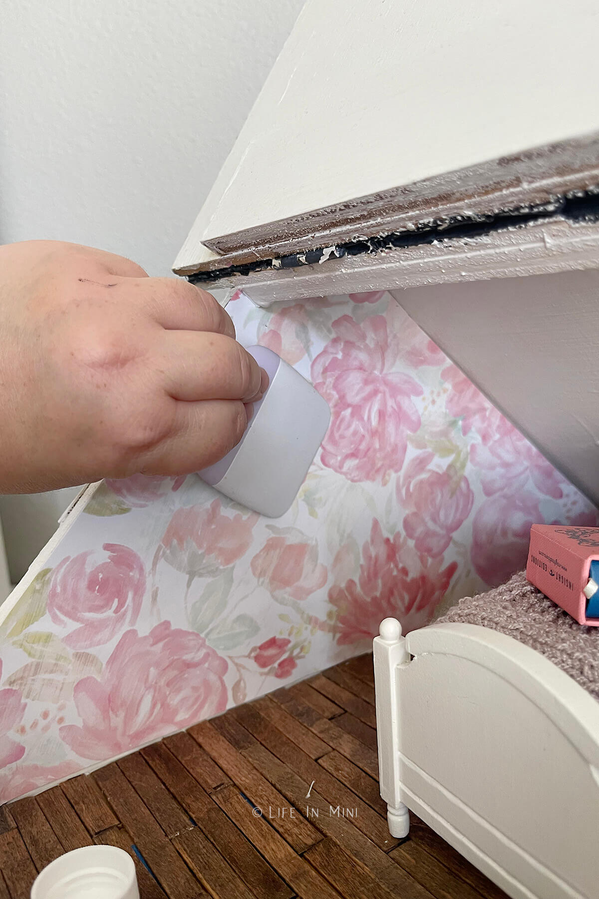 A hand smoothing out floral scrap paper used as wall paper in a dollhouse