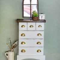 A white dollhouse dresser with a wood top and feet and brass drawer pulls with a mirror and accessories on it