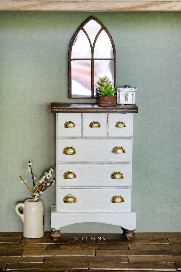 A white dollhouse dresser with a wood top and feet and brass drawer pulls with a mirror and accessories on it