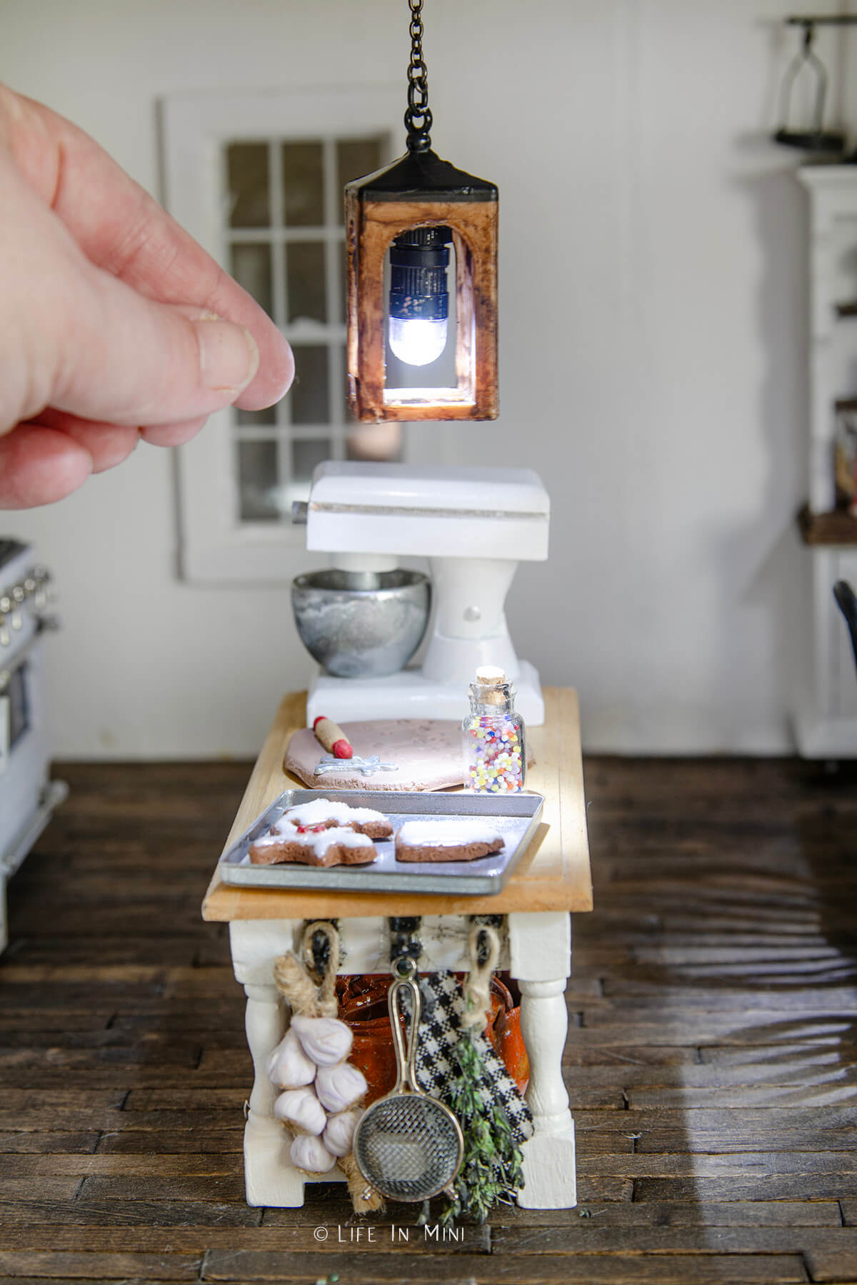 A hand placing a miniature lantern pendant light lit and hanging in a dollhouse kitchen