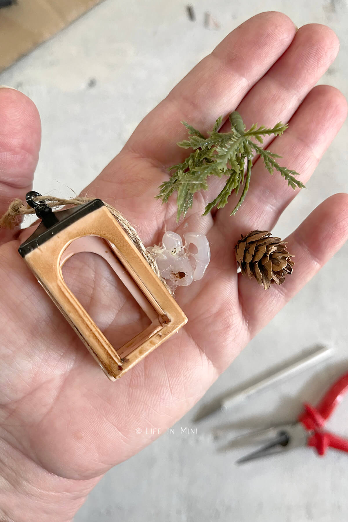 A hand holding a miniature lantern ornament with the greenery, pinecone and excess glue removed