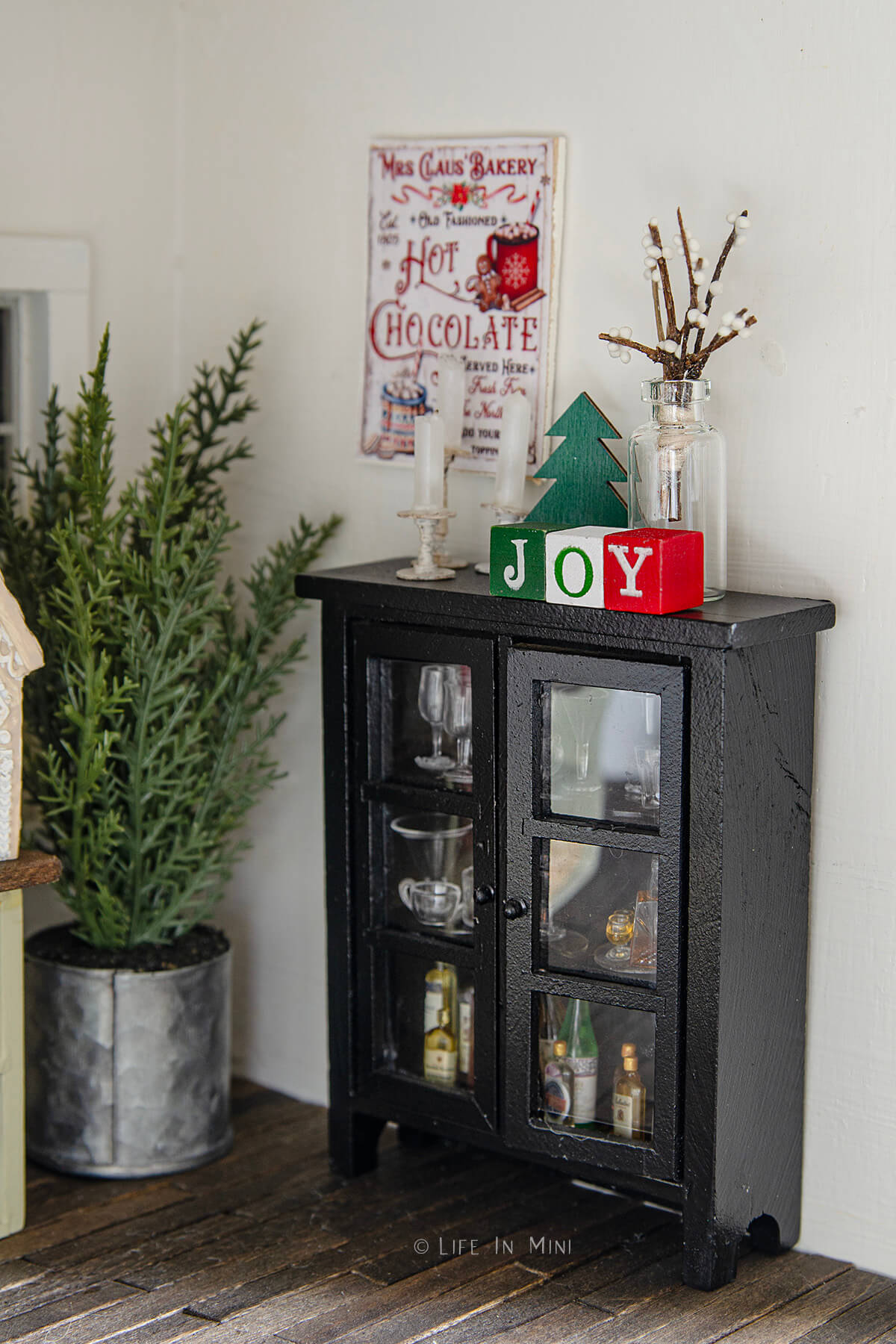 A small dollhouse cabinet filled with mini glassware and bottles, decorated for Christmas