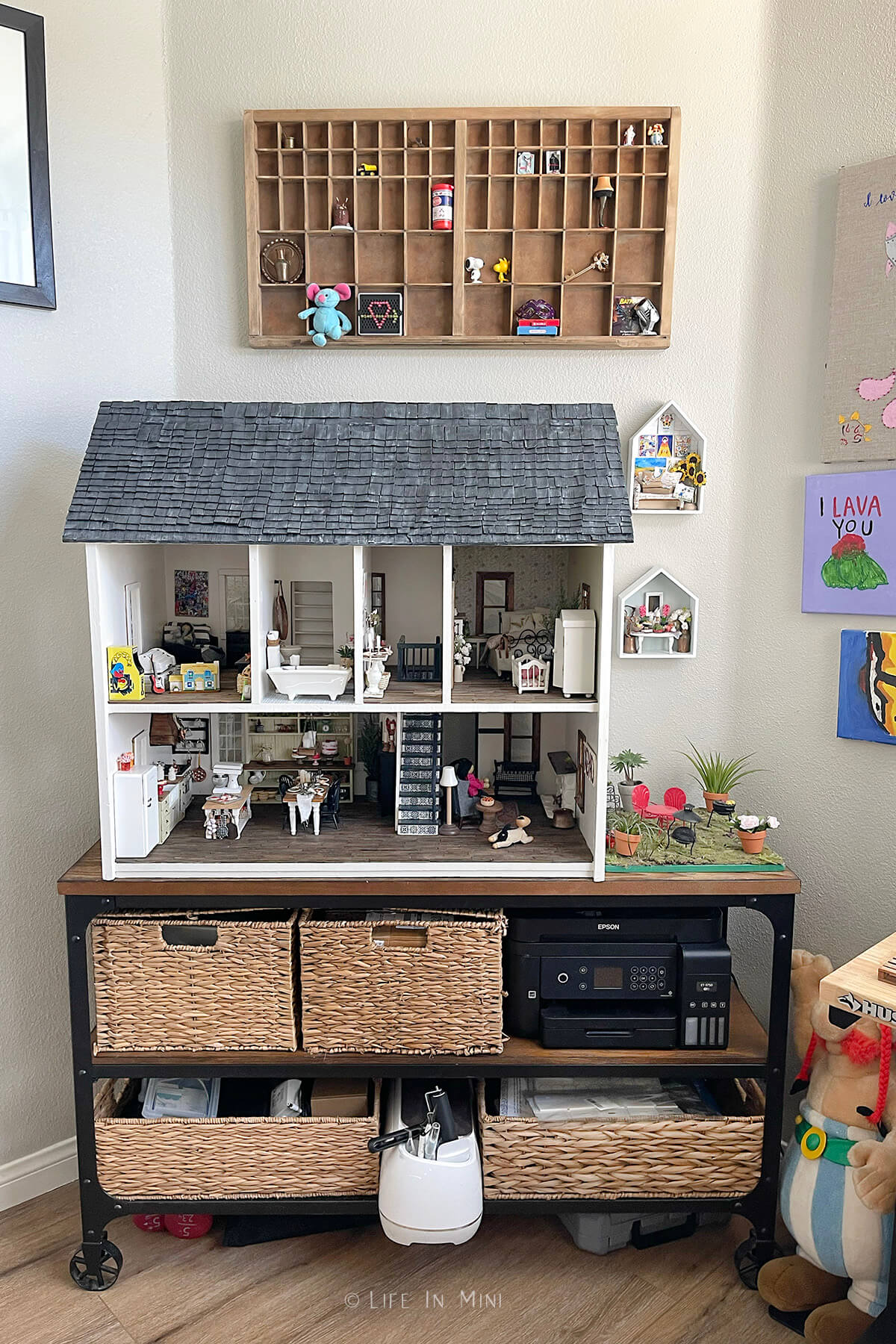 A dollhouse sitting on a rolling cart with baskets in it with a wooden printers drawer hanging over it