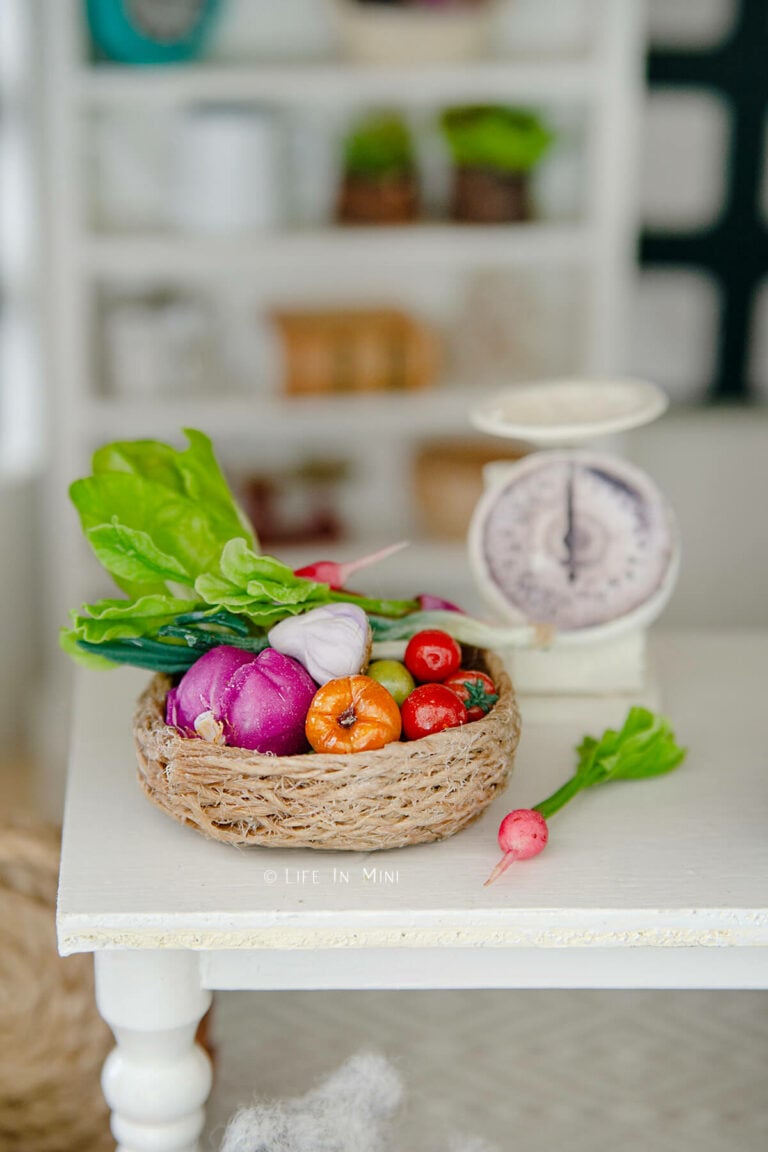 A flat miniature basket with assorted mini clay vegetables in it and kitchen scale behind it