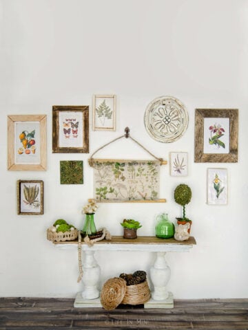 A miniature scene with a white and wood console table with various green and natural wood items on it and various botanical prints on the wall