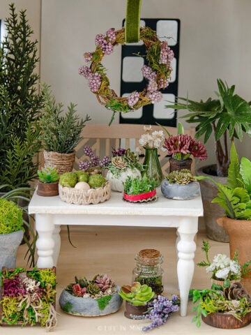 A white table with various miniature plants, a grapevine wreath hanging from above and even more plants below the table -all made from various materials.