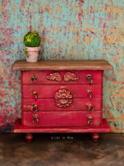 Closeup of a dollhouse dresser painted red with gold antique finish and reliefs in a rustic scene.