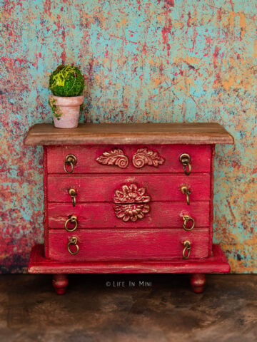 Closeup of a dollhouse dresser painted red with gold antique finish and reliefs in a rustic scene.