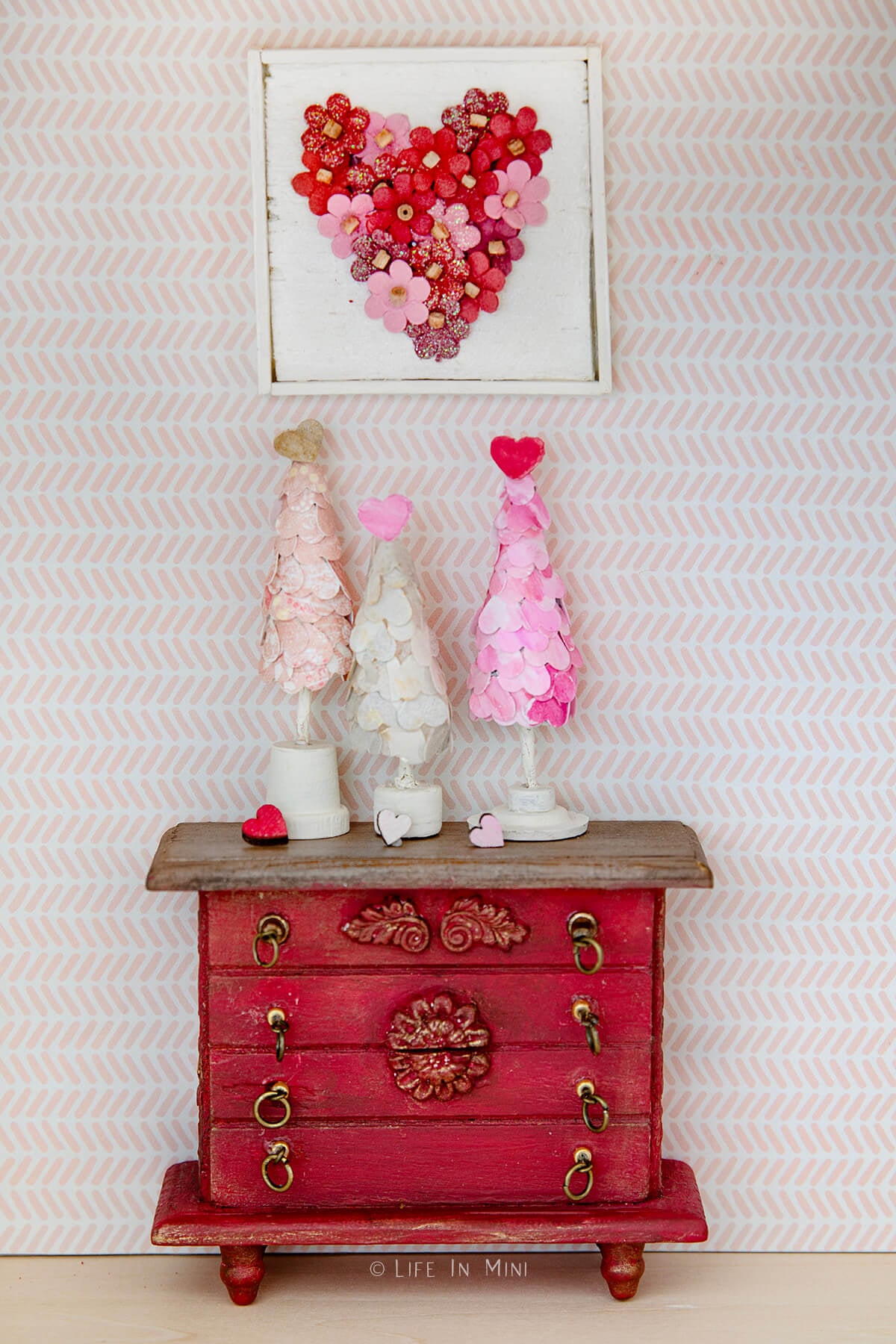 Closeup of three mini heart valentine trees sitting on top a red dollhouse dresser with a framed heart made of mini flowers over it.