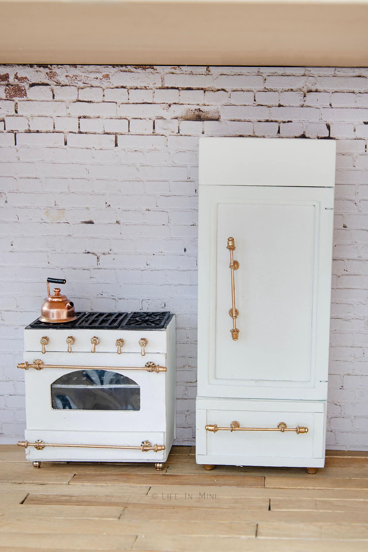 A white dollhouse refrigerator next to a white dollhouse oven both with gold handles on it.