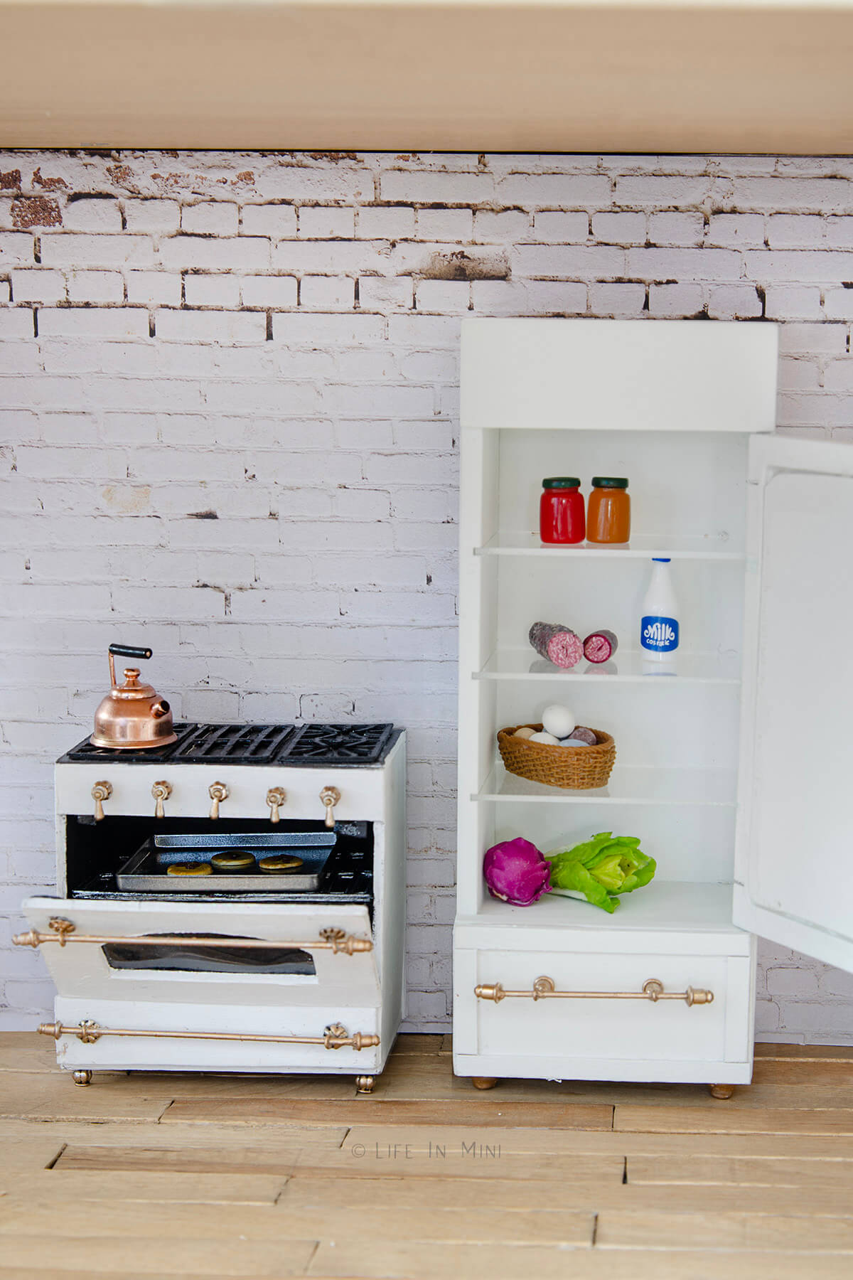 A white dollhouse refrigerator with the door open and miniature groceries in it next to a white dollhouse oven with door open and cookies in it.