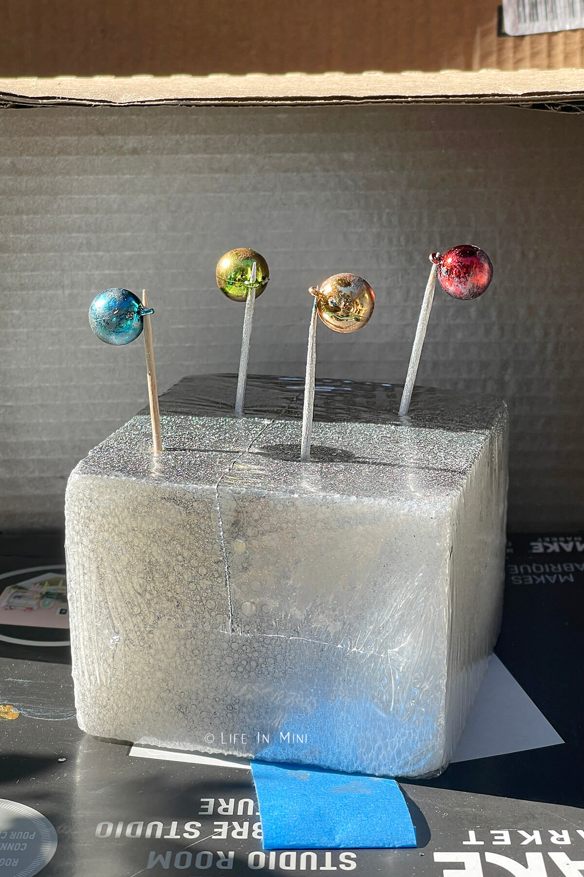 Mini Christmas ornaments propped with toothpicks stuck in a block of styrofoam getting spray painted.