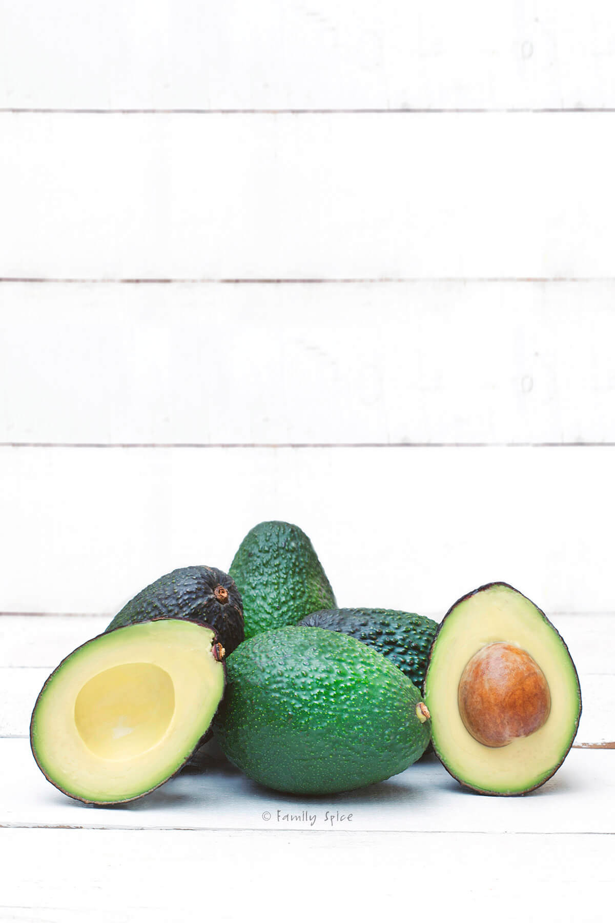 Several avocados, some cut in half, on a white wood background.